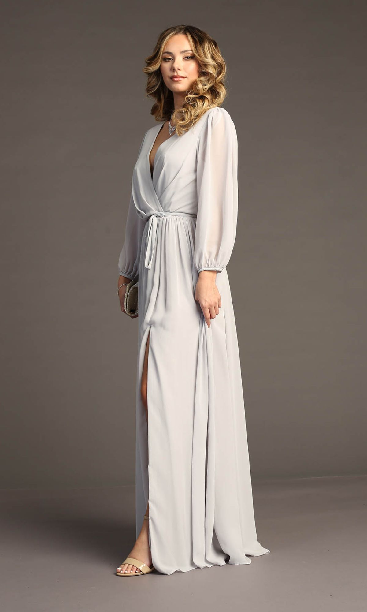 A woman wears the Long-Sleeve Long Formal Dress: Chicas J012B, a full-length light gray gown with long sleeves and a front slit, while holding a small clutch.